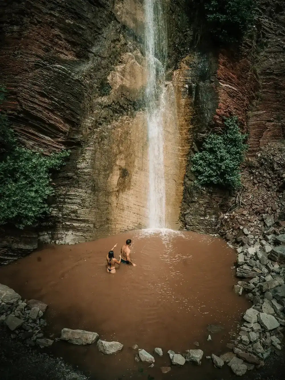 Two explorers wading beneath a towering waterfall